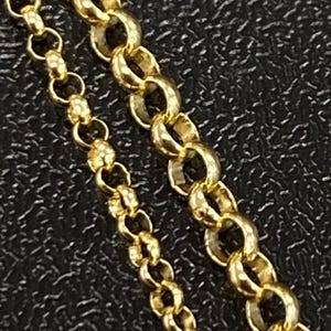 May include: Close-up of two gold necklaces with different chain styles. One chain features small, round links, while the other has larger, interlocking oval links. The necklaces are set against a dark, textured background.