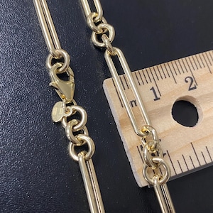 May include: Close-up of a gold-colored chain necklace with a lobster clasp. The chain features a combination of elongated links and circular links. A wooden ruler is placed next to the chain to measure its length.