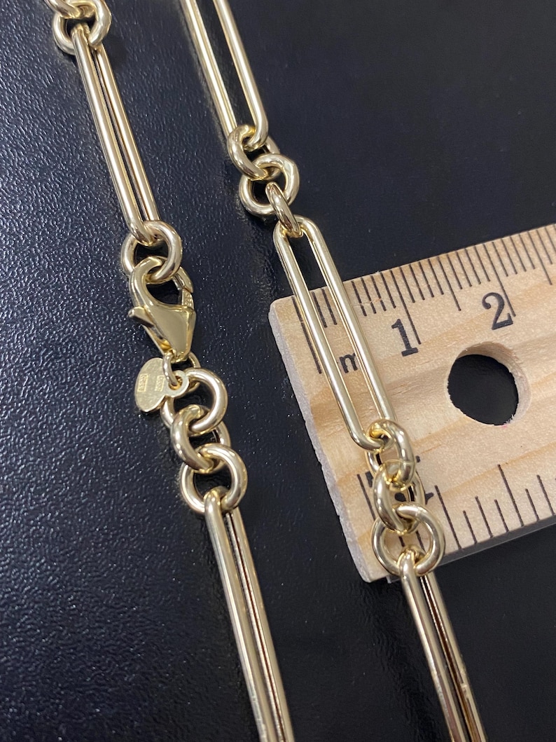 May include: Close-up of a gold-colored chain necklace with a lobster clasp. The chain features a combination of elongated links and circular links. A wooden ruler is placed next to the chain to measure its length.