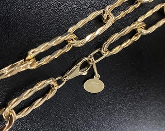 May include: Close-up of a gold-colored chain necklace with a twisted rope design. The necklace has large, elongated links and a lobster clasp closure. A small, oval charm hangs near the clasp.