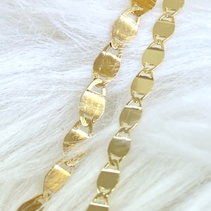 May include: Two gold chains with oval links. The chains are laying on a white fuzzy surface.