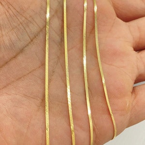 Puede incluir: Primer plano de un collar de cadena de oro con un dise&ntilde;o plano y estrecho. La cadena se muestra sobre una mano, con varios hilos de la cadena visibles.