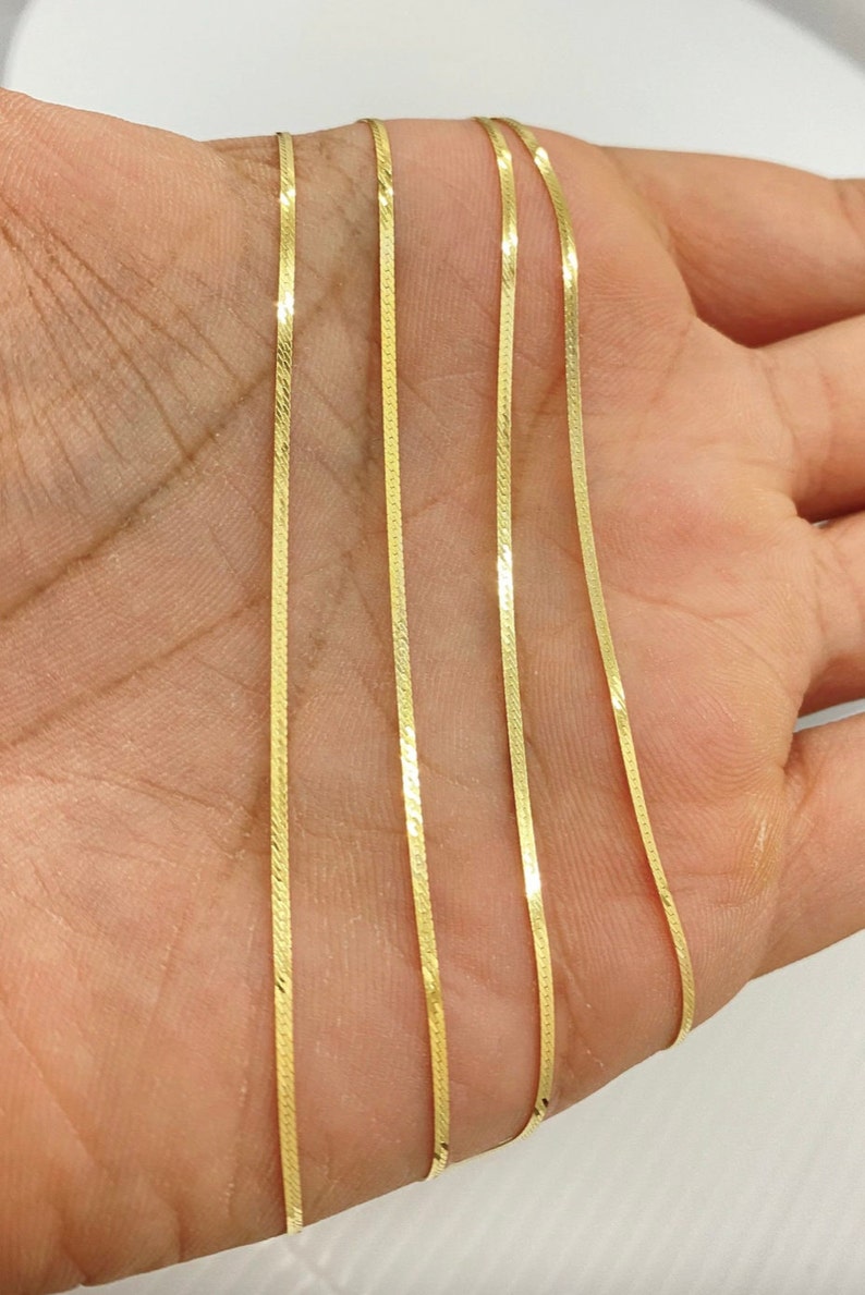 Puede incluir: Primer plano de un collar de cadena de oro con un dise&ntilde;o plano y estrecho. La cadena se muestra sobre una mano, con varios hilos de la cadena visibles.