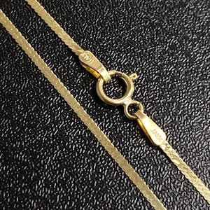 May include: Close-up of a gold herringbone chain necklace with a lobster clasp. The necklace is made of small, flat, interlocking links. The clasp is marked "ITALY". The chain is displayed against a textured black background.