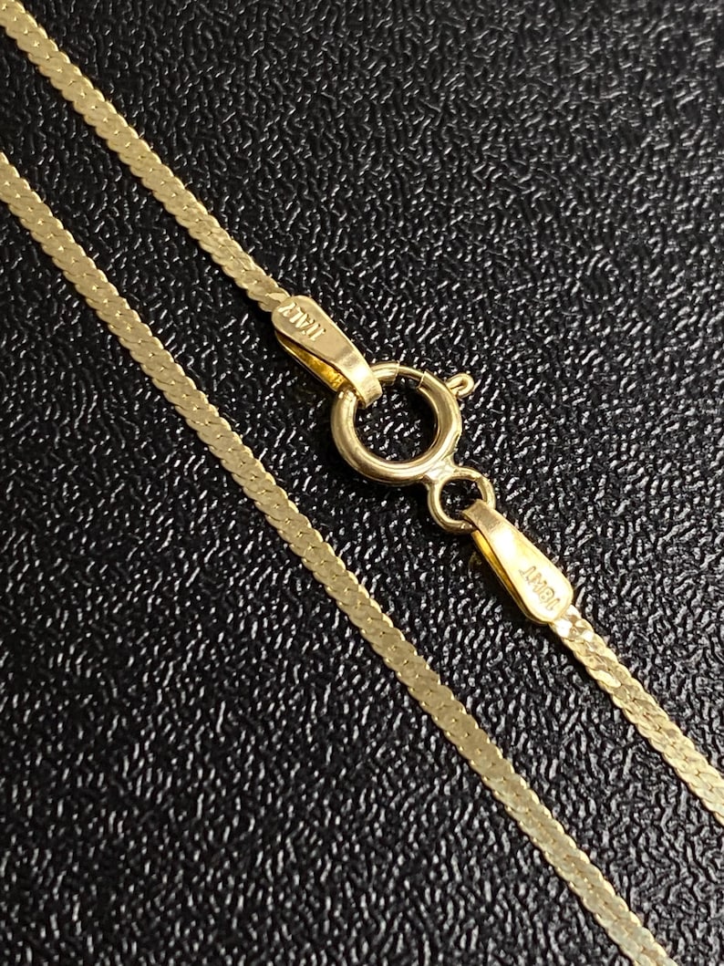 May include: Close-up of a gold herringbone chain necklace with a lobster clasp. The necklace is made of small, flat, interlocking links. The clasp is marked "ITALY". The chain is displayed against a textured black background.