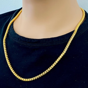 May include: A gold chain necklace with a box chain design. The chain is worn by a person with a black shirt.
