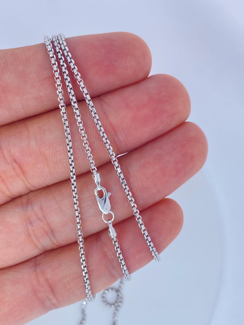 May include: A silver-colored box chain necklace with a lobster clasp. The chain is held in a hand against a white background. The chain is made of small, square links.