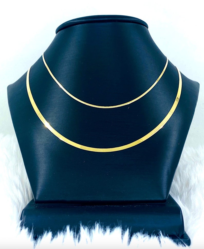 Puede incluir: Dos collares de cadena de oro en un expositor negro. Las cadenas est&aacute;n superpuestas, con la cadena m&aacute;s gruesa en la parte inferior.