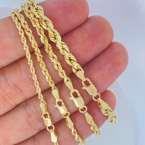 Può includere: Quattro collane a catena a corda color oro di vari spessori. Ogni catena ha una chiusura a moschettone. Le catene sono esposte su uno sfondo chiaro.