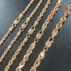 May include: Close-up of five gold-colored rope chains with lobster claw clasps. The chains vary in thickness and are displayed against a dark background. Each clasp is detailed with a small design.