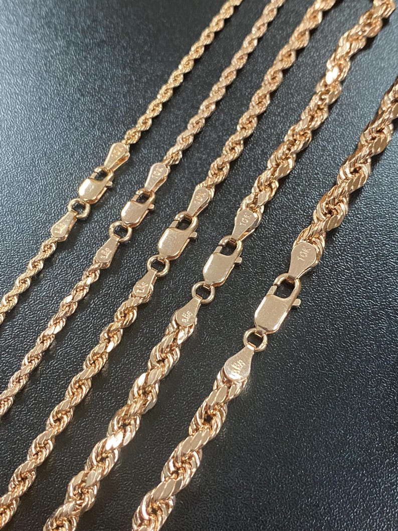 May include: Close-up of five gold-colored rope chains with lobster claw clasps. The chains vary in thickness and are displayed against a dark background. Each clasp is detailed with a small design.