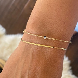 May include: Two gold bracelets are shown. The top bracelet is a delicate chain with a small blue and white evil eye charm. The bottom bracelet is a flat, textured band. The bracelets are worn on a wrist.