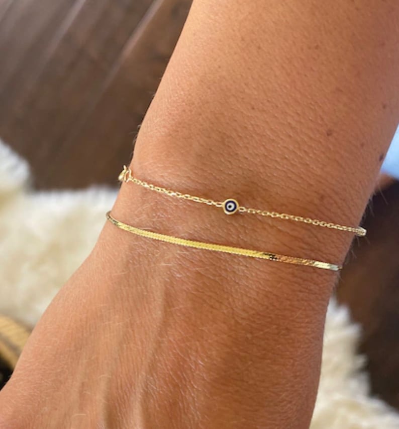 May include: Two gold bracelets are shown. The top bracelet is a delicate chain with a small blue and white evil eye charm. The bottom bracelet is a flat, textured band. The bracelets are worn on a wrist.
