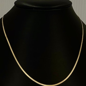 Puede incluir: Un collar de cadena de oro en un expositor negro. La cadena es fina y tiene un acabado liso y brillante.