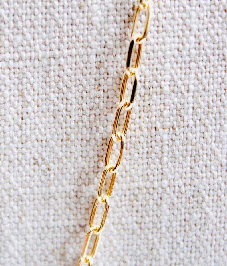 May include: A gold chain necklace with oval links. The chain is lying on a white fabric background.