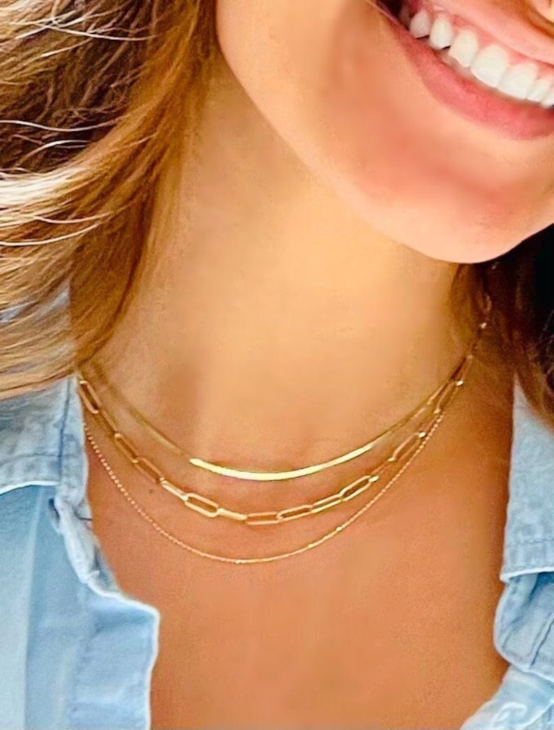May include: A layered gold necklace set. The necklaces include a delicate chain, a chain with rectangular links, and a curved bar pendant. The jewelry is worn over a light blue denim shirt.