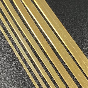Puede incluir: Un conjunto de eslabones de cadena planos de color dorado dispuestos en diagonal sobre una superficie negra. Las cadenas son de diferentes anchos.