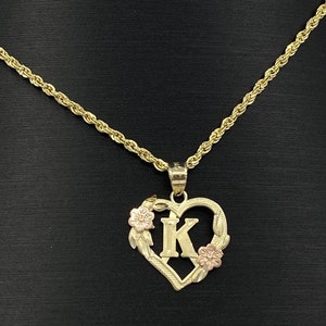 Puede incluir: Un collar de oro con un colgante en forma de coraz&oacute;n con un dise&ntilde;o floral y la letra "K" en el centro. El collar est&aacute; en una cadena de cuerda de oro.