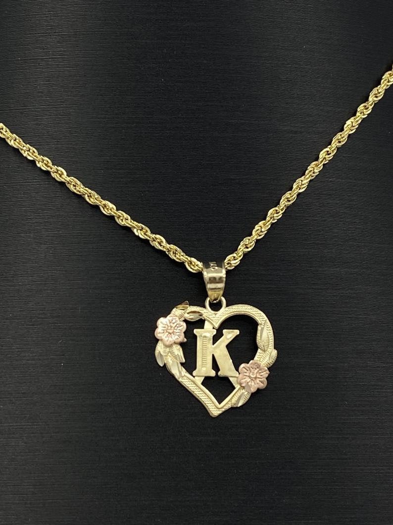 Puede incluir: Un collar de oro con un colgante en forma de coraz&oacute;n con un dise&ntilde;o floral y la letra "K" en el centro. El collar est&aacute; en una cadena de cuerda de oro.