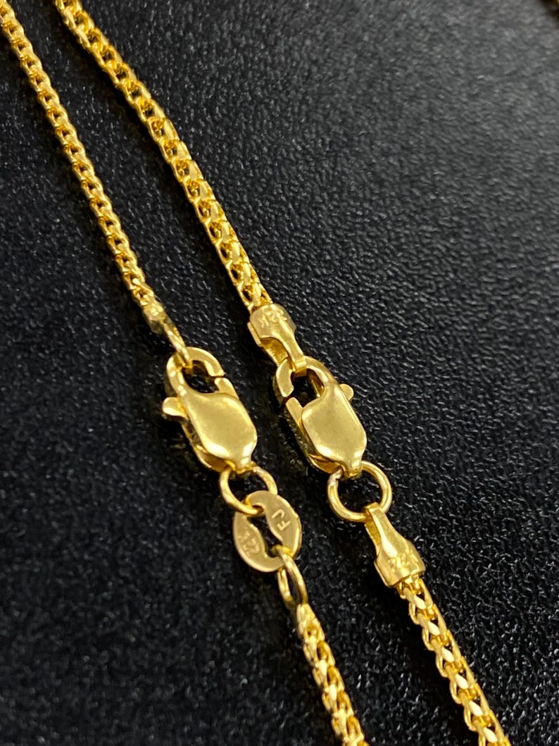 May include: Close-up view of two gold chains with lobster clasps. The chains are delicate and feature a box chain design.  The clasps are marked with '750', indicating 18k gold. The chains are displayed against a dark background.