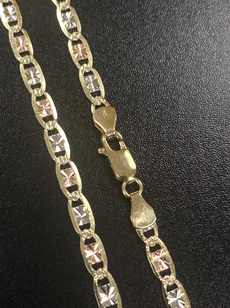 May include: Gold-tone necklace with a unique design. The chain features alternating oval links with a three-tone starburst pattern. The clasp is also gold-toned. The necklace is displayed on a dark surface.