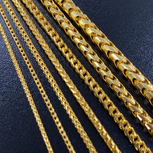 Puede incluir: Primer plano de varias cadenas de collar doradas. Las cadenas varían en grosor y diseño, algunas presentan un patrón tejido y otras eslabones rectangulares más sólidos. Las cadenas se muestran sobre un fondo oscuro, resaltando su brillo metálico.