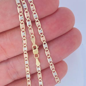 K&ouml;nnte beinhalten: Eine zarte Halskette mit dreifarbiger Kette mit abwechselnd goldenen, ros&eacute;goldenen und silberfarbenen Gliedern. Die Kette wird in einer Hand gehalten, wodurch ihr filigranes Design und die warmen Farbt&ouml;ne des Metalls zur Geltung kommen. Der Verschluss ist sichtbar.