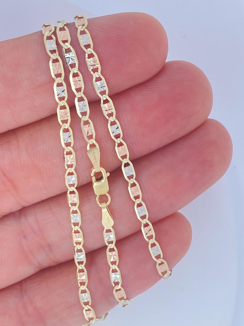 K&ouml;nnte beinhalten: Eine zarte Halskette mit dreifarbiger Kette mit abwechselnd goldenen, ros&eacute;goldenen und silberfarbenen Gliedern. Die Kette wird in einer Hand gehalten, wodurch ihr filigranes Design und die warmen Farbt&ouml;ne des Metalls zur Geltung kommen. Der Verschluss ist sichtbar.