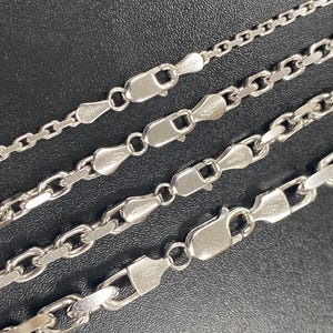 May include: Four silver-colored chain necklaces with different link styles and clasp closures. The chains are displayed against a dark background, showcasing their varying designs and textures. The necklaces are likely made of sterling silver.