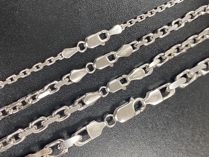 May include: Four silver-colored chain necklaces with different link styles and clasp closures. The chains are displayed against a dark background, showcasing their varying designs and textures. The necklaces are likely made of sterling silver.