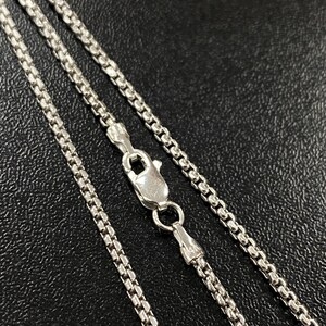May include: Close-up of a silver-colored box chain necklace with a lobster clasp. The chain is made of small, square links, and the clasp is securely fastened. The necklace is displayed against a dark background.