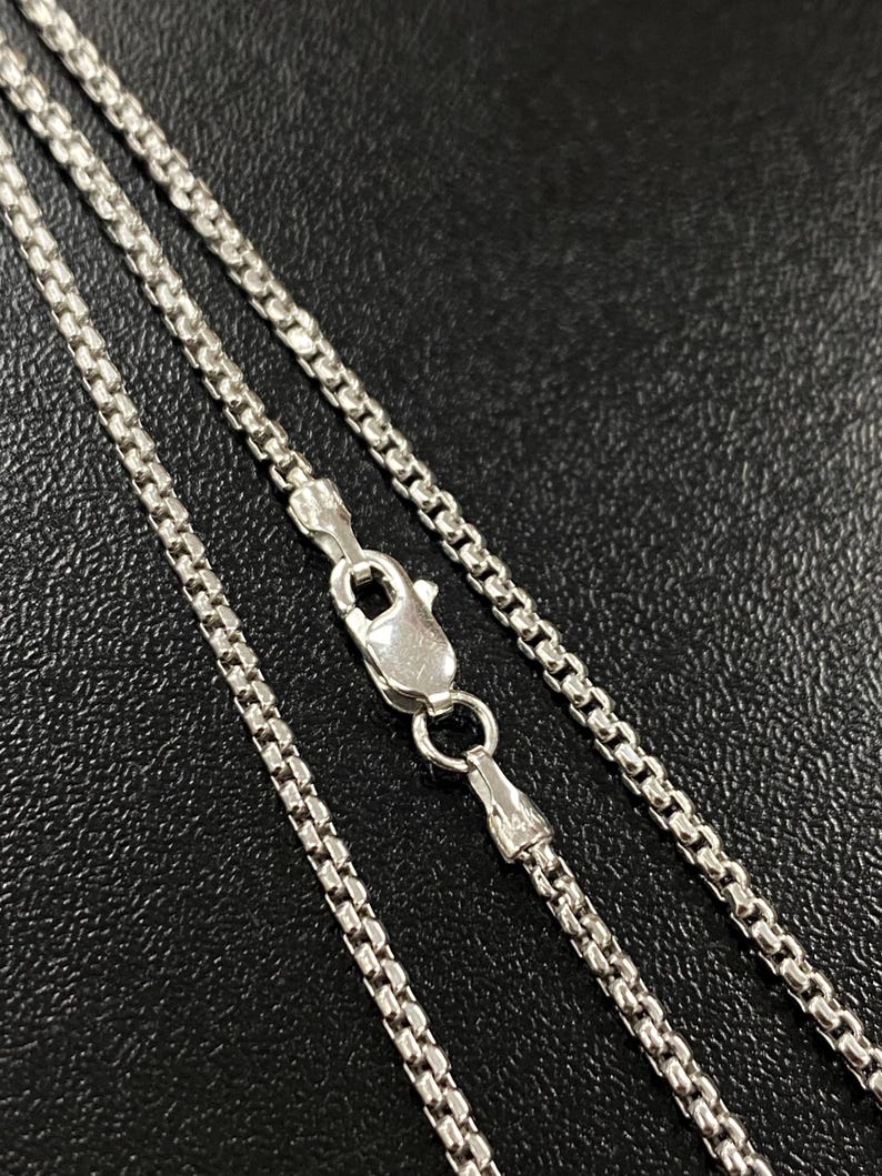 May include: Close-up of a silver-colored box chain necklace with a lobster clasp. The chain is made of small, square links, and the clasp is securely fastened. The necklace is displayed against a dark background.