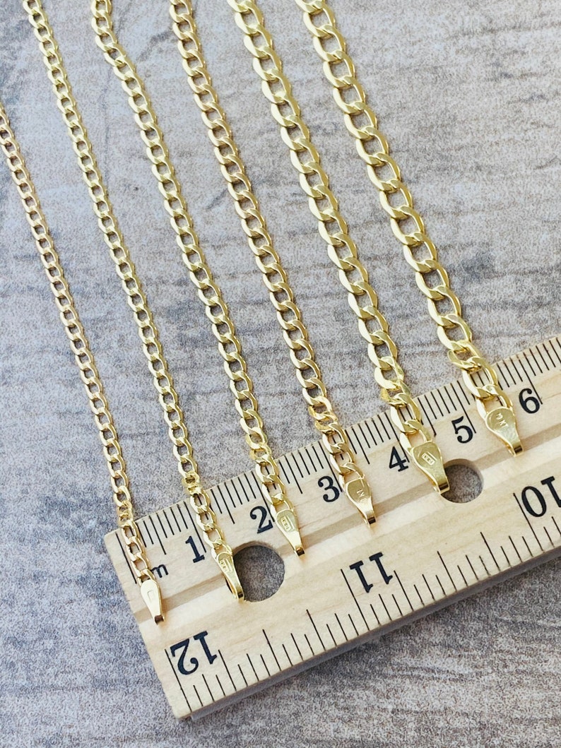 May include: Five gold chains of different lengths laid out on a wooden ruler. The chains are all the same style, with a curb link design. The ruler is marked in inches.
