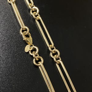 May include: Close-up of a gold-colored chain necklace. The necklace features a series of elongated, rectangular links connected by smaller, circular links. A lobster clasp is visible at the end. The background is black.