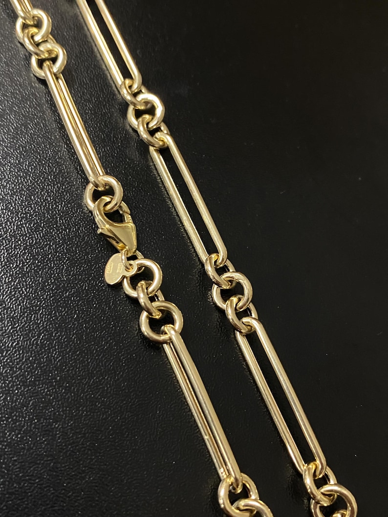 May include: Close-up of a gold-colored chain necklace. The necklace features a series of elongated, rectangular links connected by smaller, circular links. A lobster clasp is visible at the end. The background is black.