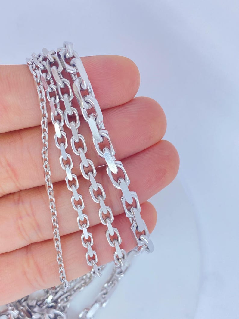 May include: A close-up of several silver-colored chain necklaces. The chains vary in style, with different link designs, including oval and rectangular shapes. The necklaces are held in a hand against a white background.