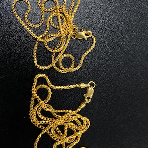 May include: Two gold-colored chain necklaces with lobster claw clasps are displayed against a dark background. The chains have a woven design, and the clasps are visible at the ends of each necklace. The chains are intertwined.
