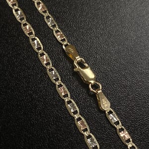 K&ouml;nnte beinhalten: Eine goldene Halskette mit Diamantschliff. Die Kette hat abwechselnd Glieder aus Gold, Ros&eacute;gold und Silber. Der Verschluss ist ebenfalls aus Gold. Die Halskette wird auf einer schwarzen Oberfl&auml;che pr&auml;sentiert.