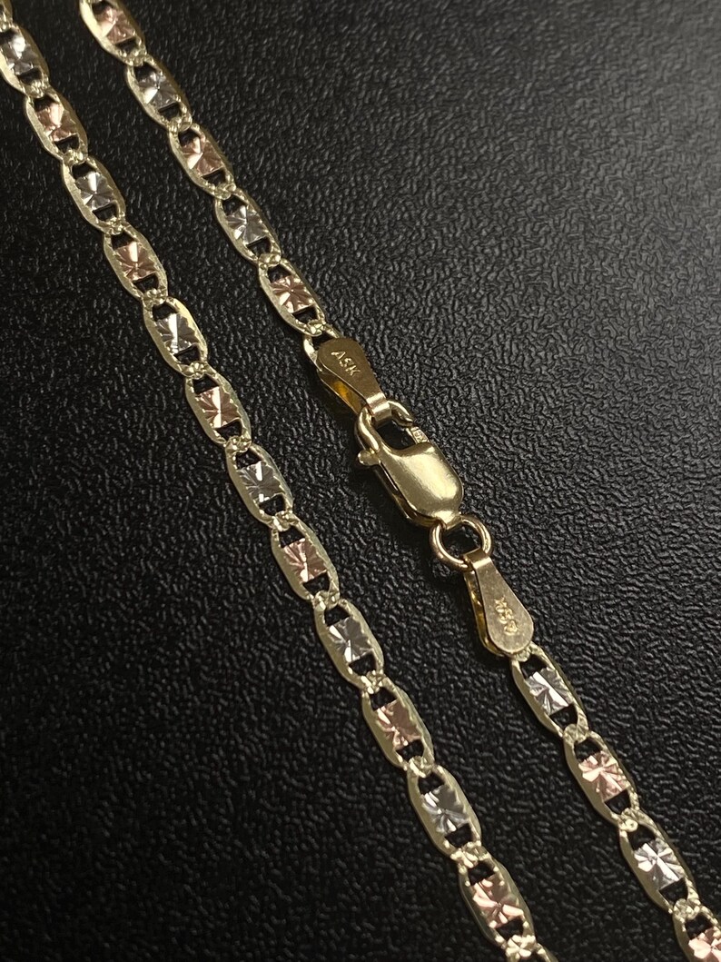 K&ouml;nnte beinhalten: Eine goldene Halskette mit Diamantschliff. Die Kette hat abwechselnd Glieder aus Gold, Ros&eacute;gold und Silber. Der Verschluss ist ebenfalls aus Gold. Die Halskette wird auf einer schwarzen Oberfl&auml;che pr&auml;sentiert.
