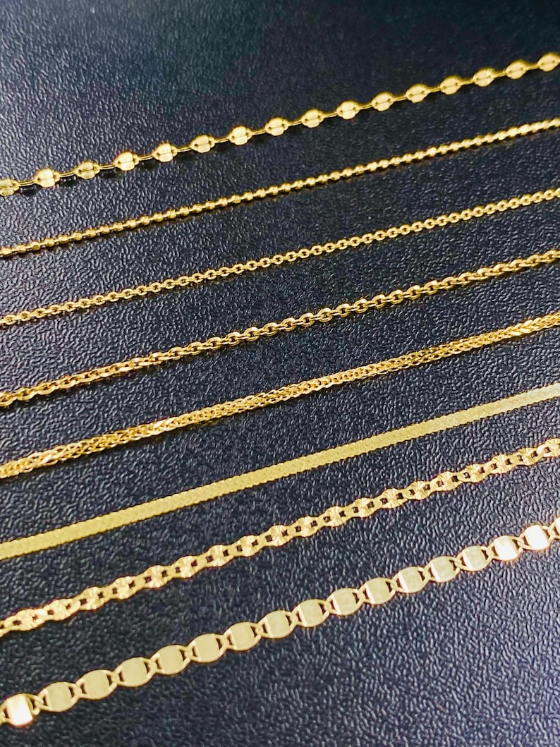 May include: A selection of gold necklaces featuring various chain designs, including beaded, cable, and flat styles. The chains are displayed on a dark surface, highlighting their golden colour and intricate details. The image shows a variety of necklace styles.