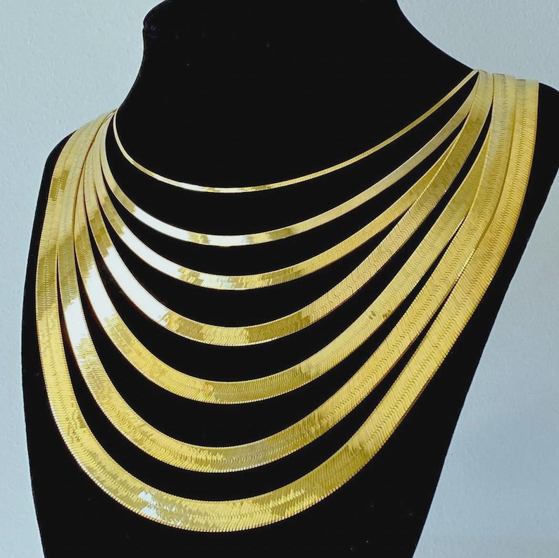 Puede incluir: Un conjunto de collares de cadena de serpiente de tono dorado de diferentes anchos, superpuestos en un expositor negro.