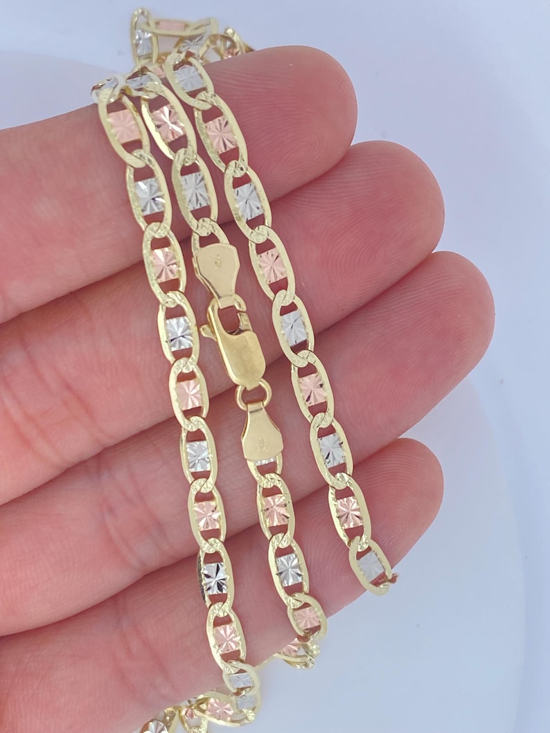 May include: A gold-toned chain necklace with alternating oval links and decorative accents in gold, rose gold, and silver. The necklace features a lobster clasp closure. The jewelry is displayed against a light background.