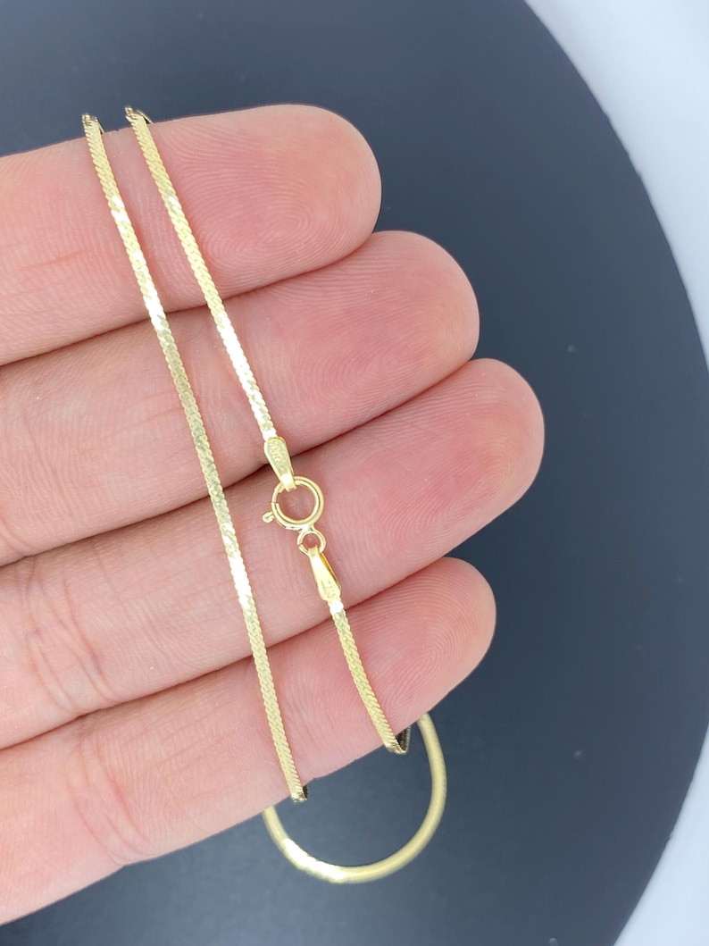 May include: A gold-colored herringbone chain necklace with a lobster clasp. The chain is thin and flat, with a textured surface that catches the light. The necklace is shown against a neutral background.