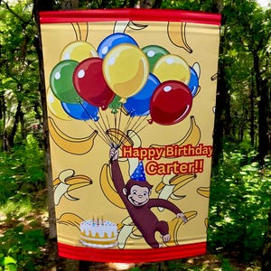 May include: A yellow banner with a cartoon monkey holding balloons. The banner has a yellow background with a pattern of bananas. The text "Happy Birthday Carter!" is printed on the banner.