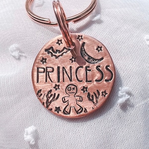 Könnte beinhalten: Ein kupferfarbener Haustieranhänger mit einer schwarzen Aufschrift "PRINCESS". Der Anhänger zeigt einen Halbmond, eine Fledermaus, einen Lebkuchenmann und Sterne.