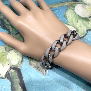 May include: A silver-toned chain bracelet with a textured, glittery finish. The bracelet features large, interlocking links and is worn on a wrist. The jewelry is displayed against a colorful, abstract background.