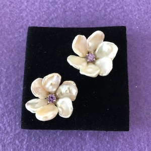 May include: A pair of flower-shaped earrings. Each earring features five off-white petals surrounding a small, round, purple gemstone. The earrings are displayed on a black velvet surface against a purple background.