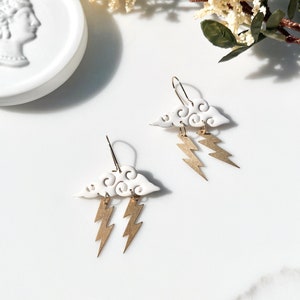 May include: White ceramic earrings shaped like clouds with gold lightning bolts hanging from them.
