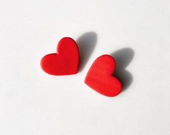 May include: Two bright red heart-shaped objects are positioned on a white surface. The hearts are solid and have a smooth texture. Shadows are cast to the side, suggesting a light source from above. The image is simple and clean.