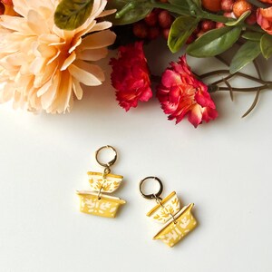 May include: A pair of gold earrings with a yellow and white patterned design. The earrings are made of ceramic and have a stacked design.